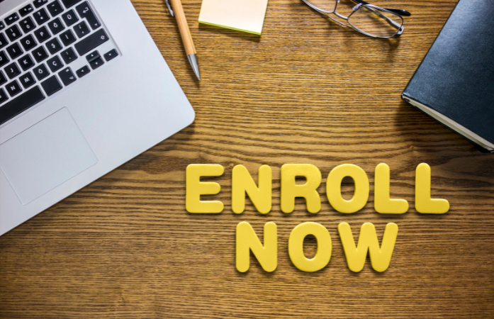  laptop, glasses, notebook and enroll now words on the screen